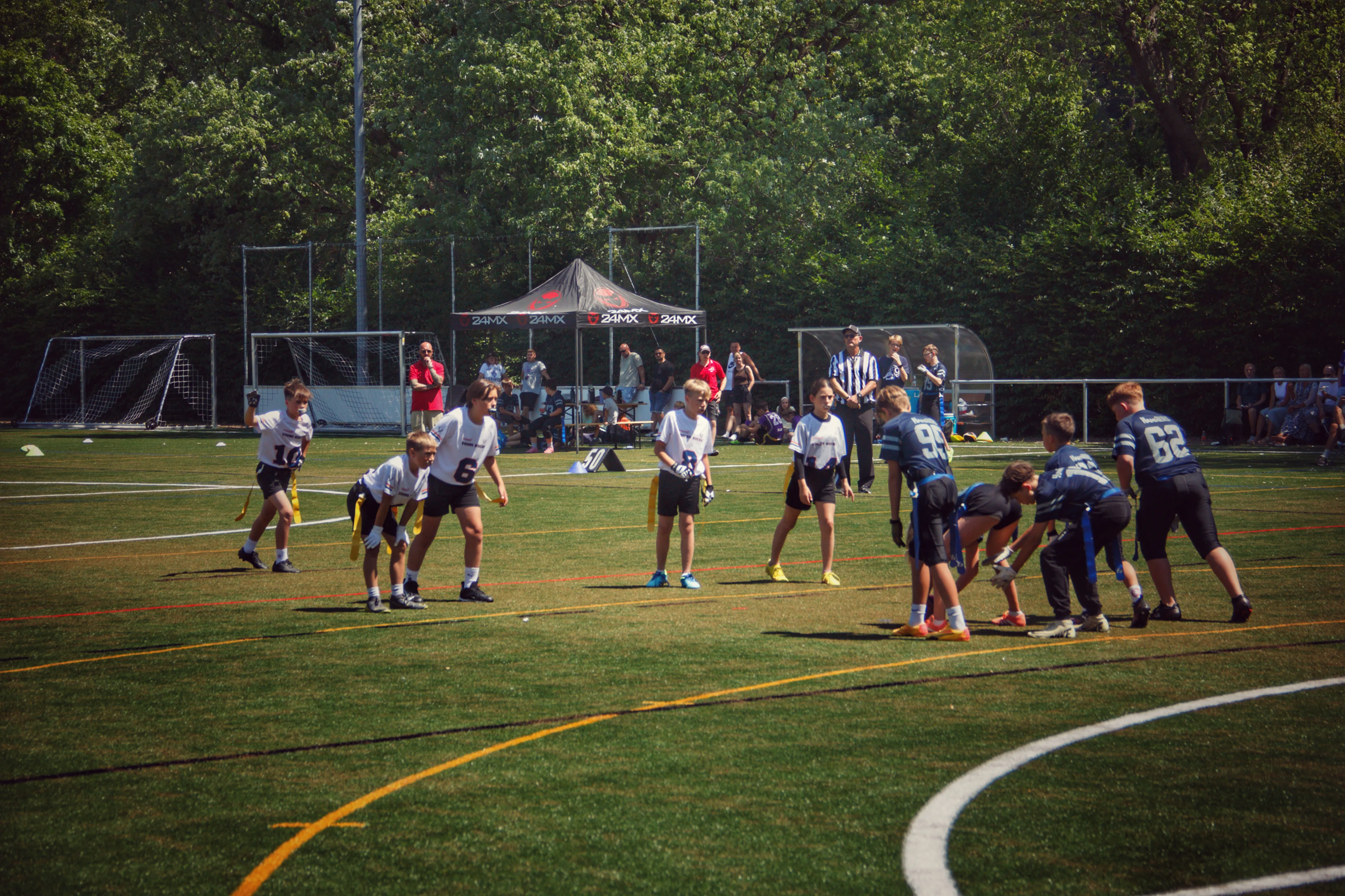 A group of players participates in a football game on a grassy field, with a pavilion and spectators in the background.
