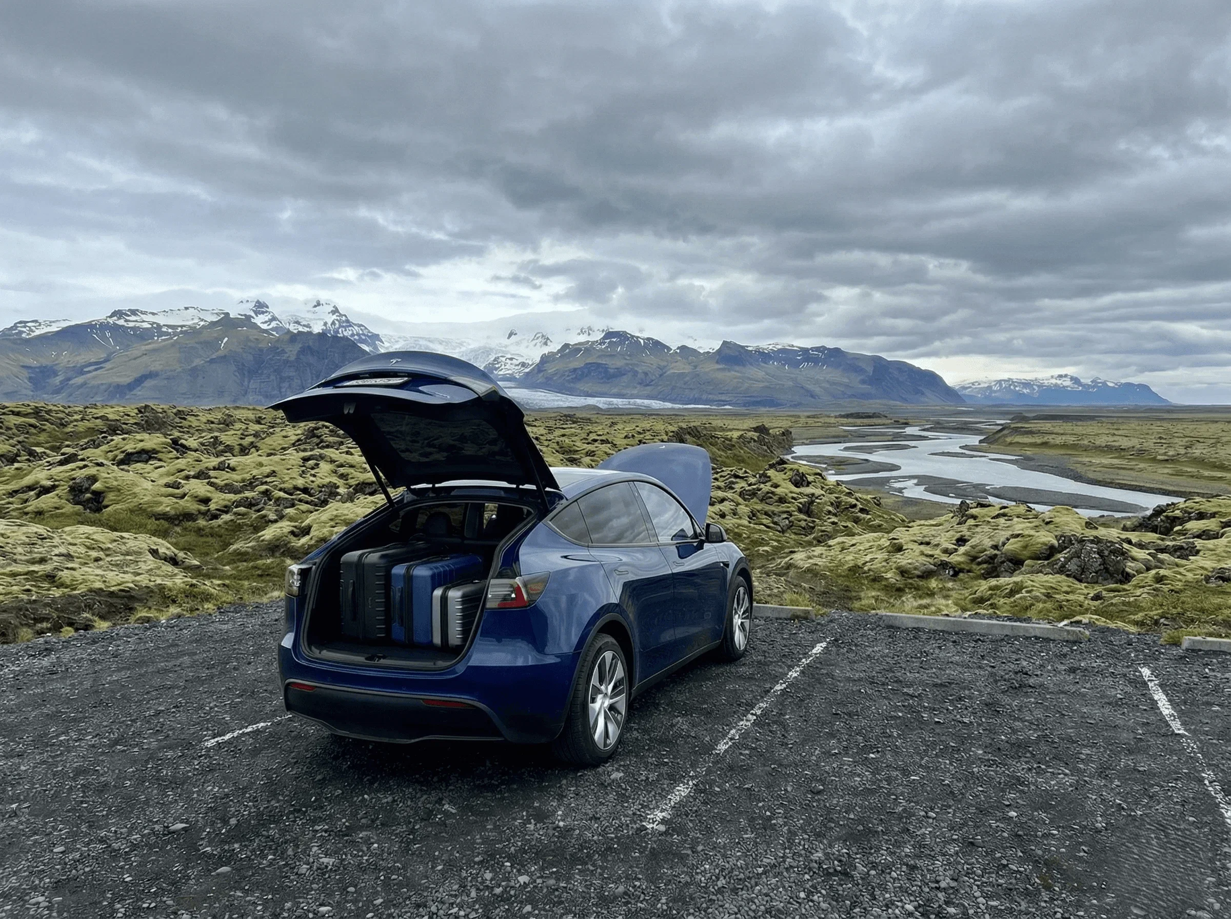 Rear view of a blue Tesla Model Y with the trunk open, packed with luggage, parked in a mossy lava field.