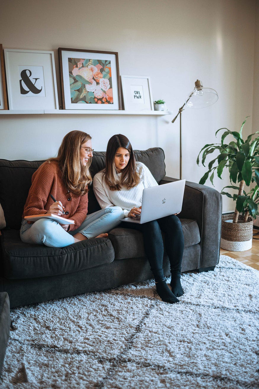 Katherine and Gabby, cofounders of CXD studio, collaborate while sitting on a gray couch next to a plant.