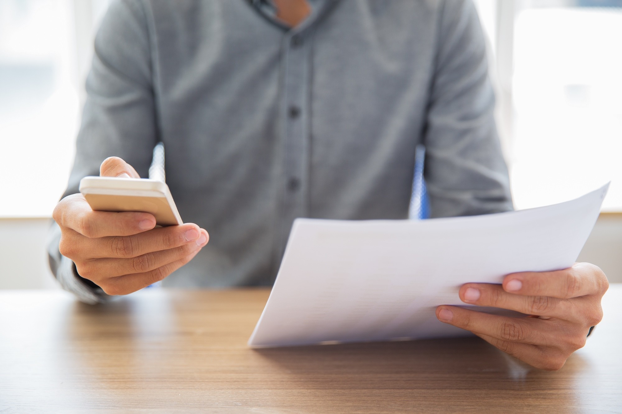 Bust of a man sitting at a desk, typing on a smartphone with his right hand and holding a sheet of paper with his left.