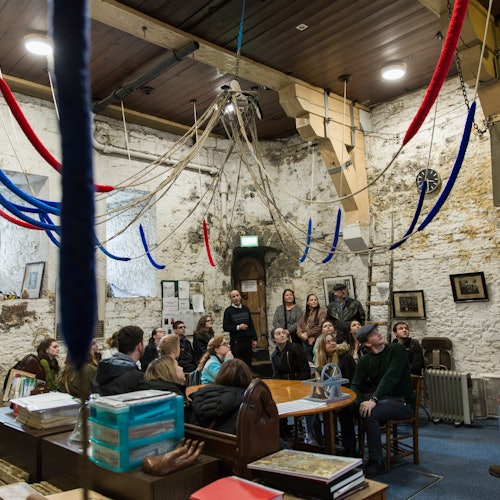 A group of people sit and stand in a rustic room with stone walls, colorful ropes hanging from the ceiling, and various framed pictures.