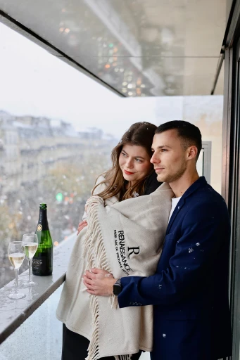 A couple stands on a city balcony wrapped in a cozy blanket, with champagne and glasses on the railing, overlooking a rainy urban landscape, creating a romantic and intimate atmosphere.