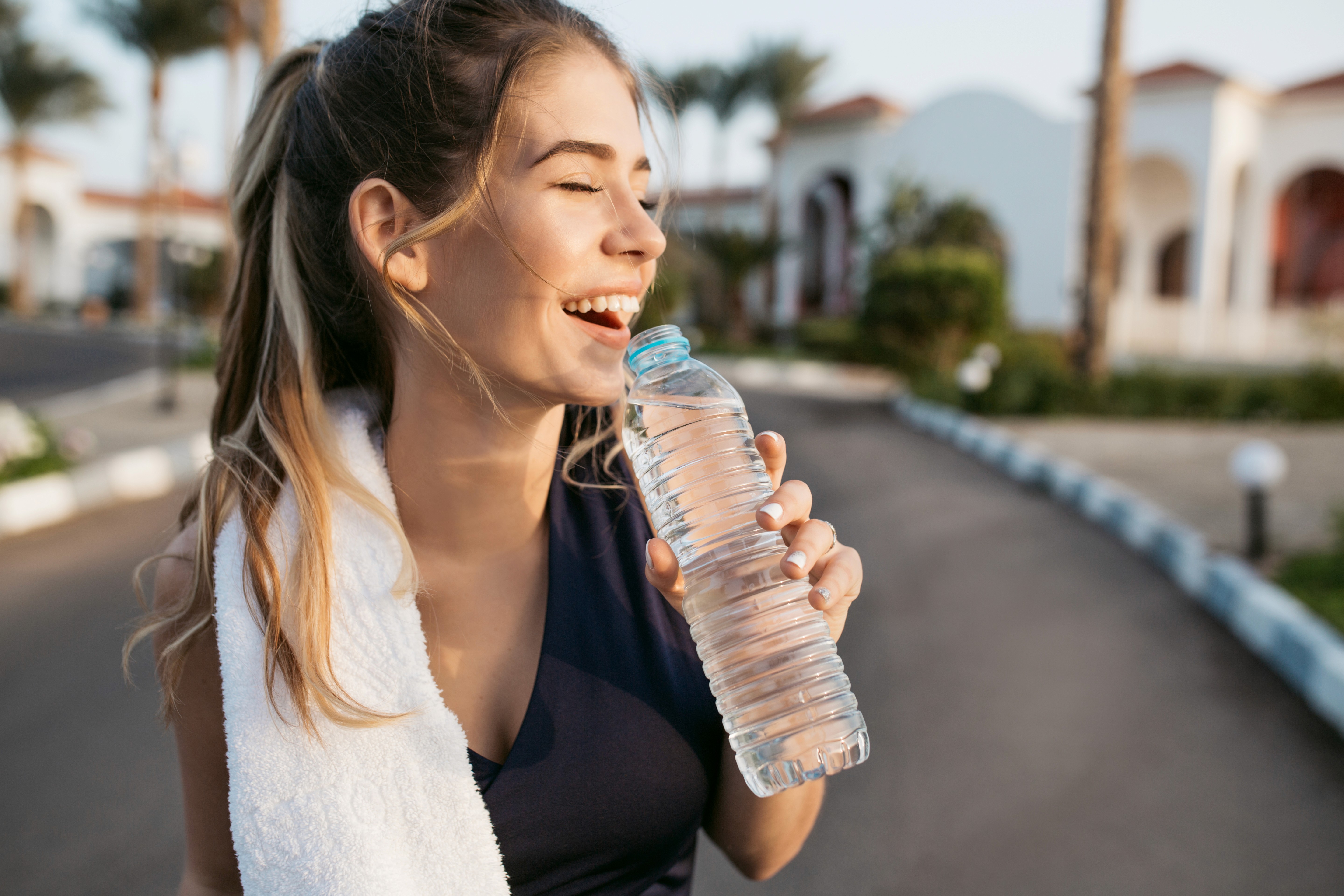 A woman with a ponytail and a towel over her shoulder holds a water bottle, smiling joyfully. The sunny outdoor setting has palm trees and buildings.