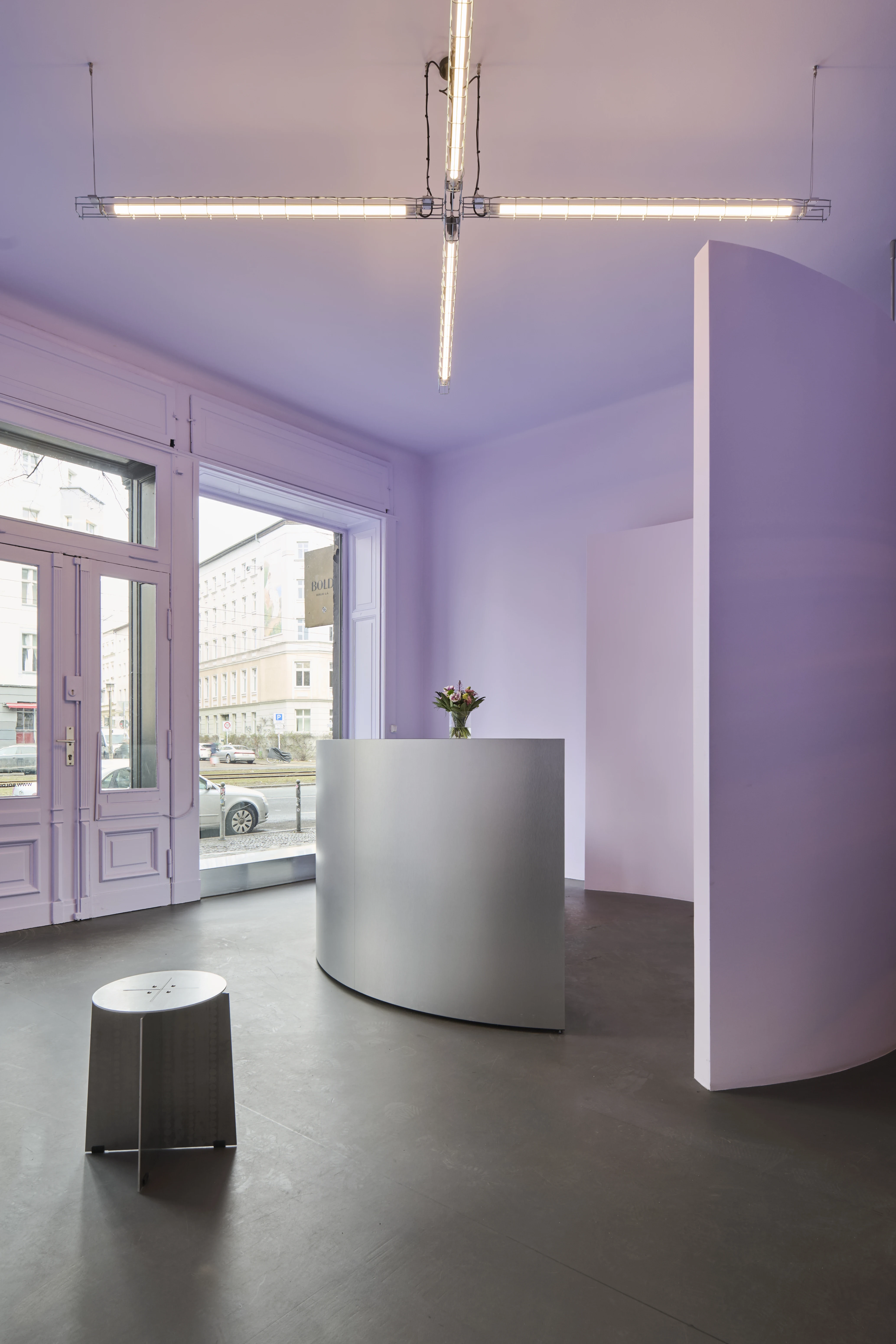 Reception area of BOLD OFFICE with curved brushed-metal desk, lavender walls, floor-to-ceiling entrance doors, industrial cross-shaped ceiling light, and a metal stool in the foreground.