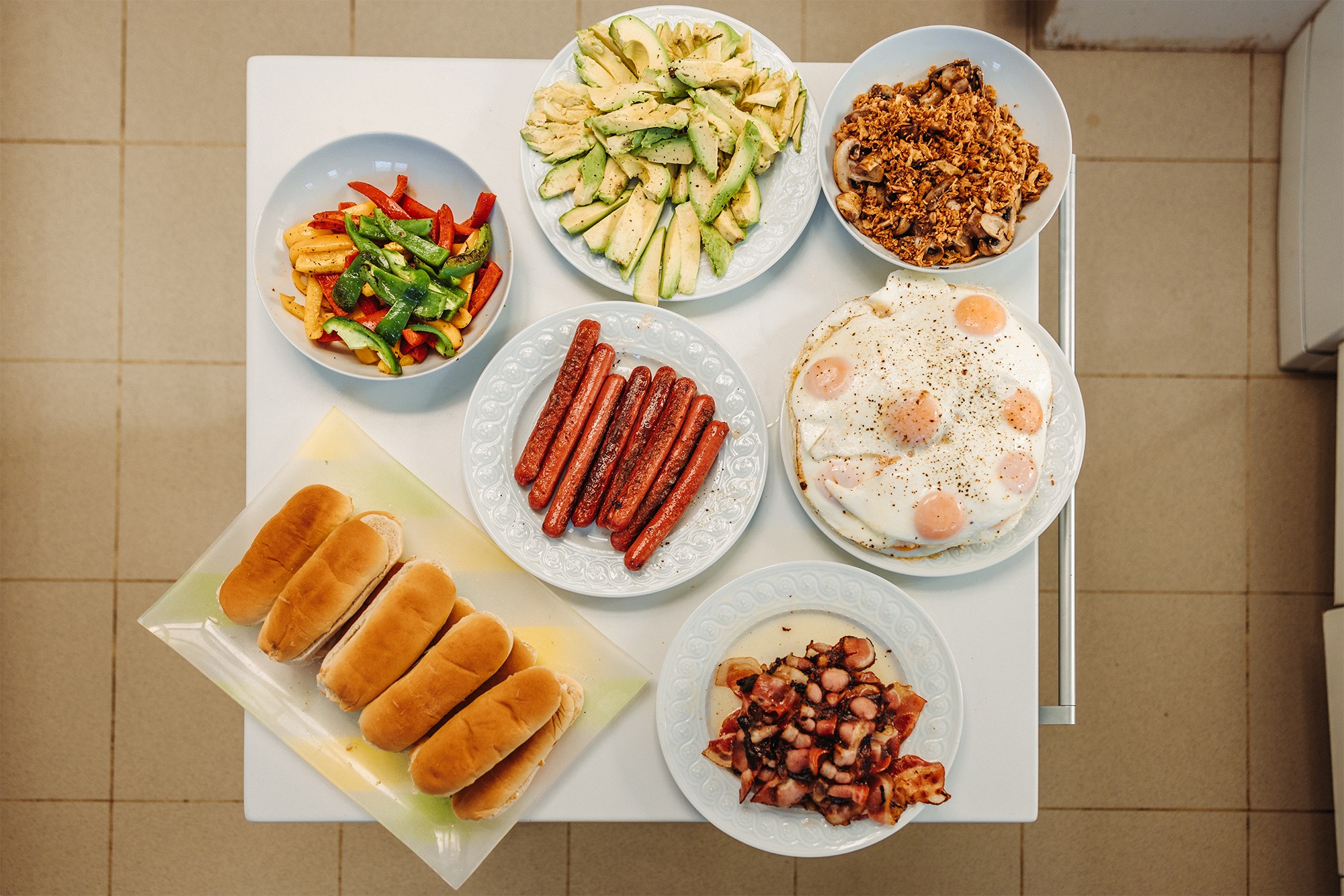 An assortment of dishes on a countertop, featuring sausages, eggs with sunny-side-up yolks, sliced avocados, grilled sausage pieces, bell peppers with potatoes, crispy onions, and hot dog buns