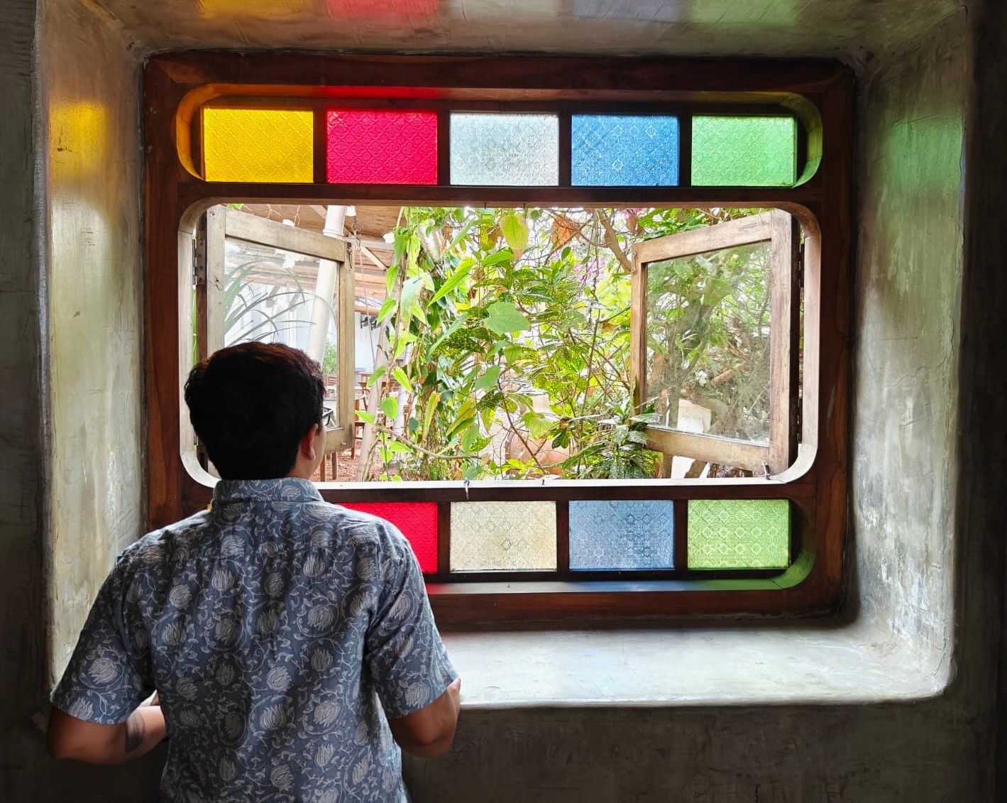 A picture of a person with short hair, wearing a floral cotton shirt, standing with their back to the camera and looking out a window. The window has a coloured glass border in yellow, red, white, blue and green, opening to a view of trees.