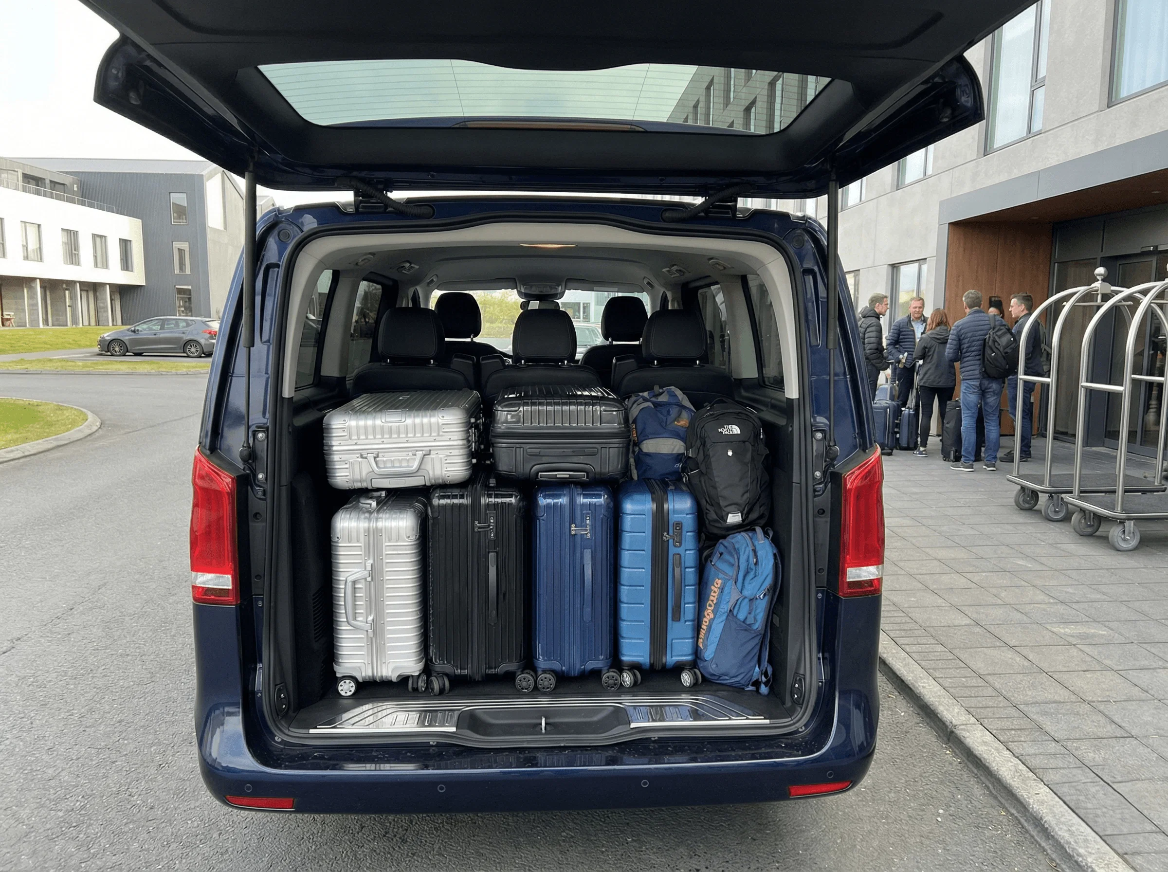 Rear view of a silver Mercedes van with the trunk open, fully packed with suitcases in a parking area.