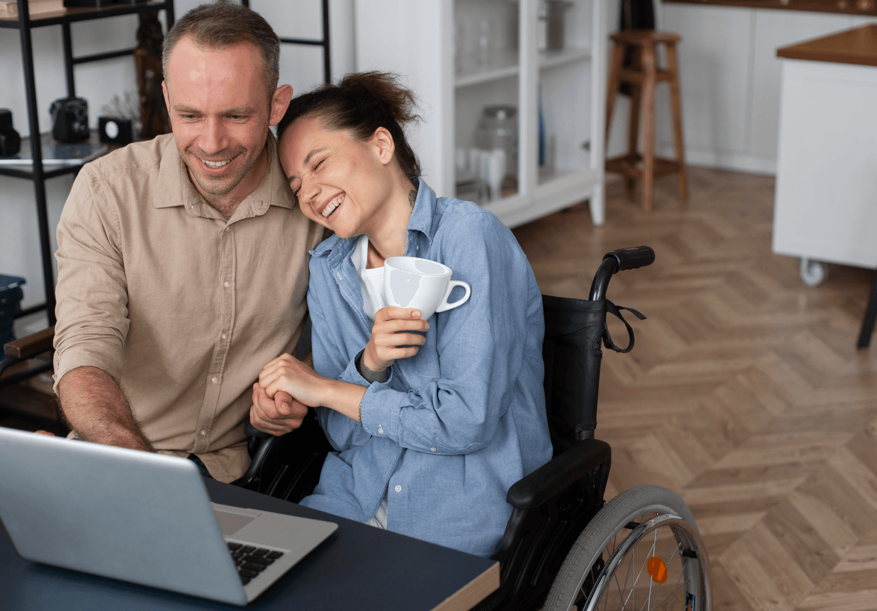 A smiling man and a woman in a wheelchair sit close together at a table, looking at a laptop. The woman holds a white mug and leans happily on the man as they share a joyful moment in a cozy room.