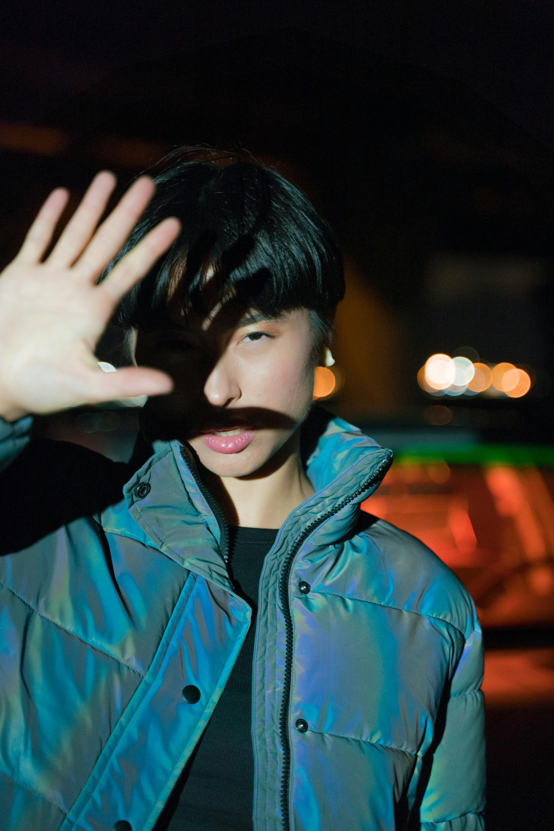 A person wearing an iridescent puffer jacket holds a hand up to shield their face from a bright light at night.