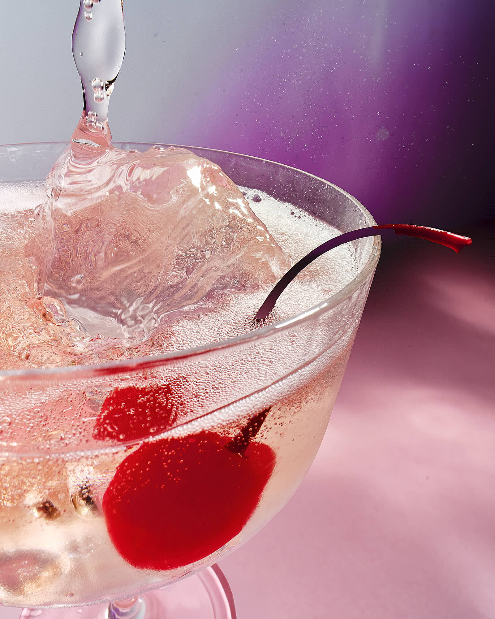 A glass of sparkling drink with a cherry and a splash of liquid against a pink and purple background.