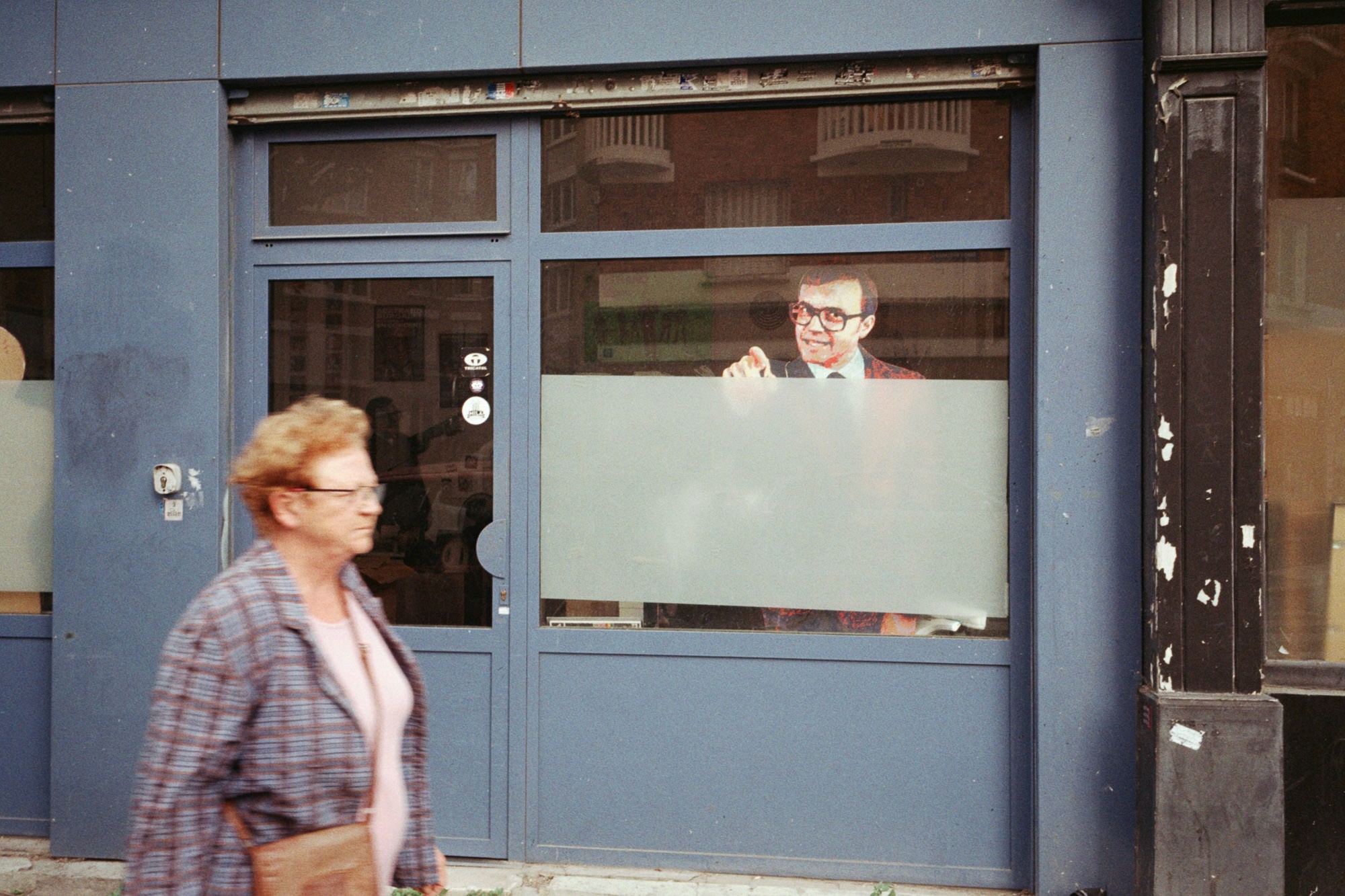 A person wearing a suit enthusiastically gestures from behind a large window of a modern building, while a casually dressed woman walks by on the sidewalk; the scene reflects urban life and business activity.