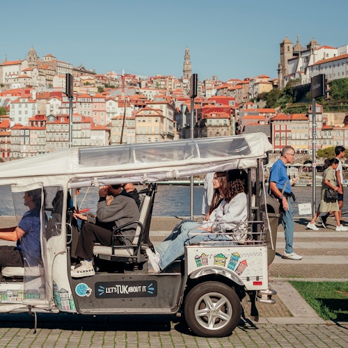 人们乘坐一辆白色三轮车靠近河畔；背景是一座城市，拥有色彩斑斓的房屋和山丘上的教堂。