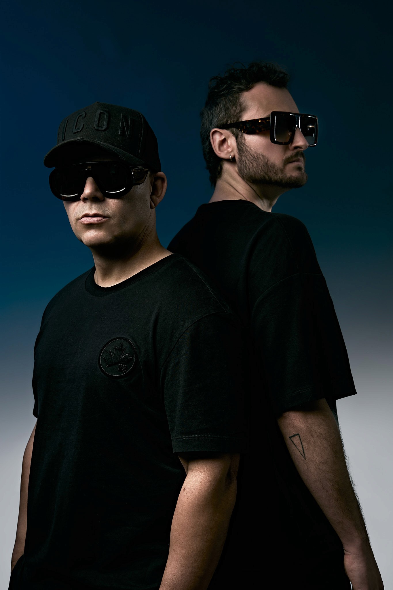 Fotografía de retrato profesional de dos hombres con gafas en estudio, con ropa negra y un fondo azul oscuro, representando moda y autenticidad con un toque de sofisticación en Palma, Mallorca.