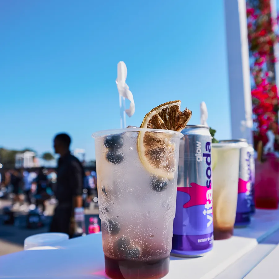 A glass of Vita Soda Glow over ice with lemon and blueberries, the cans behind it