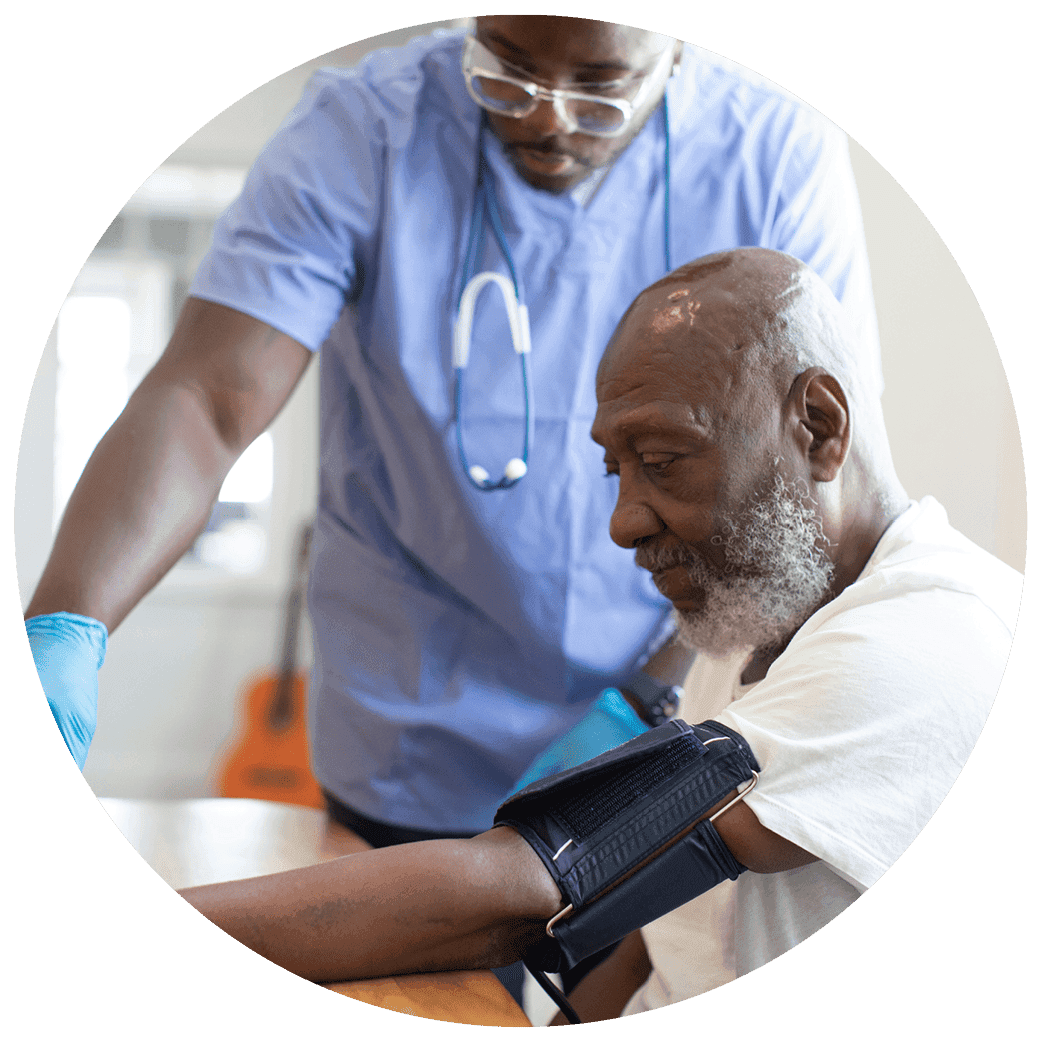 Healthcare worker wearing scrubs and gloves taking a seated person’s blood pressure using a cuff on the arm.