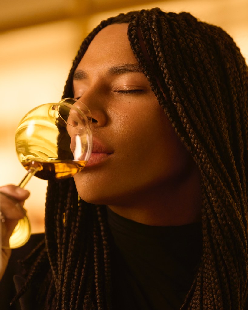 A person with braided hair gently holds a glass of whisky, savoring the aroma with their eyes closed, set against a softly lit background.
