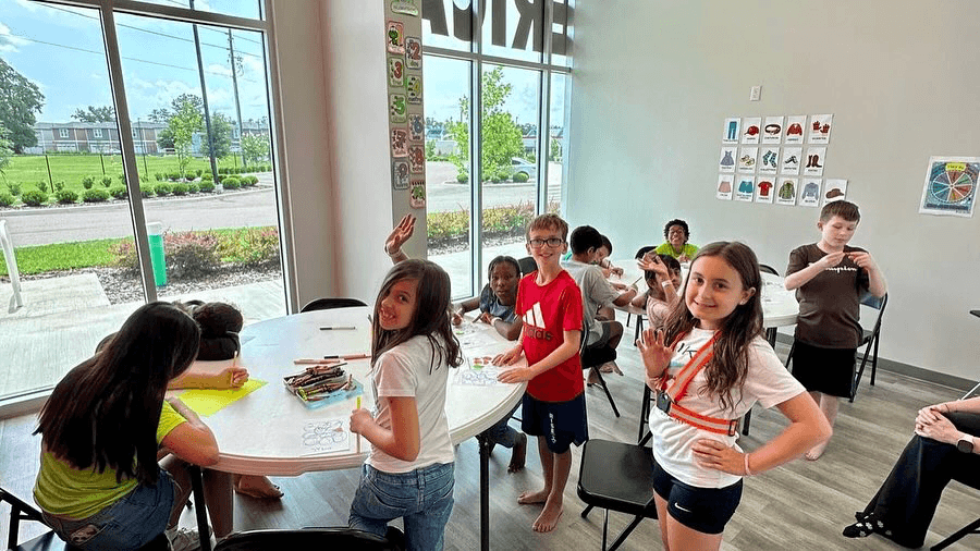 Students gathered at a tables and doing crafts, waving and smiling at the camera. 