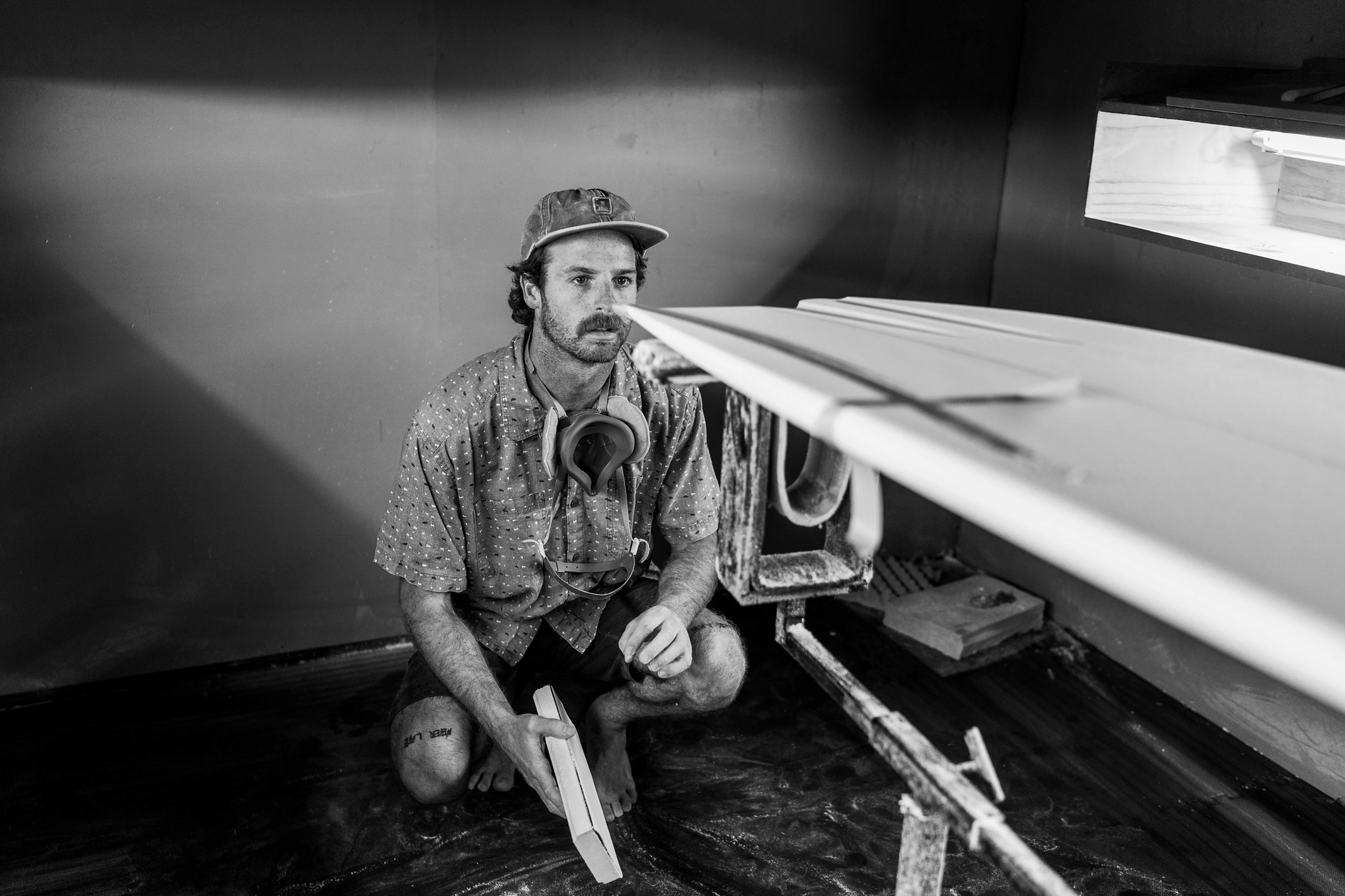 Man crouches next to surfboard in workshop setting.