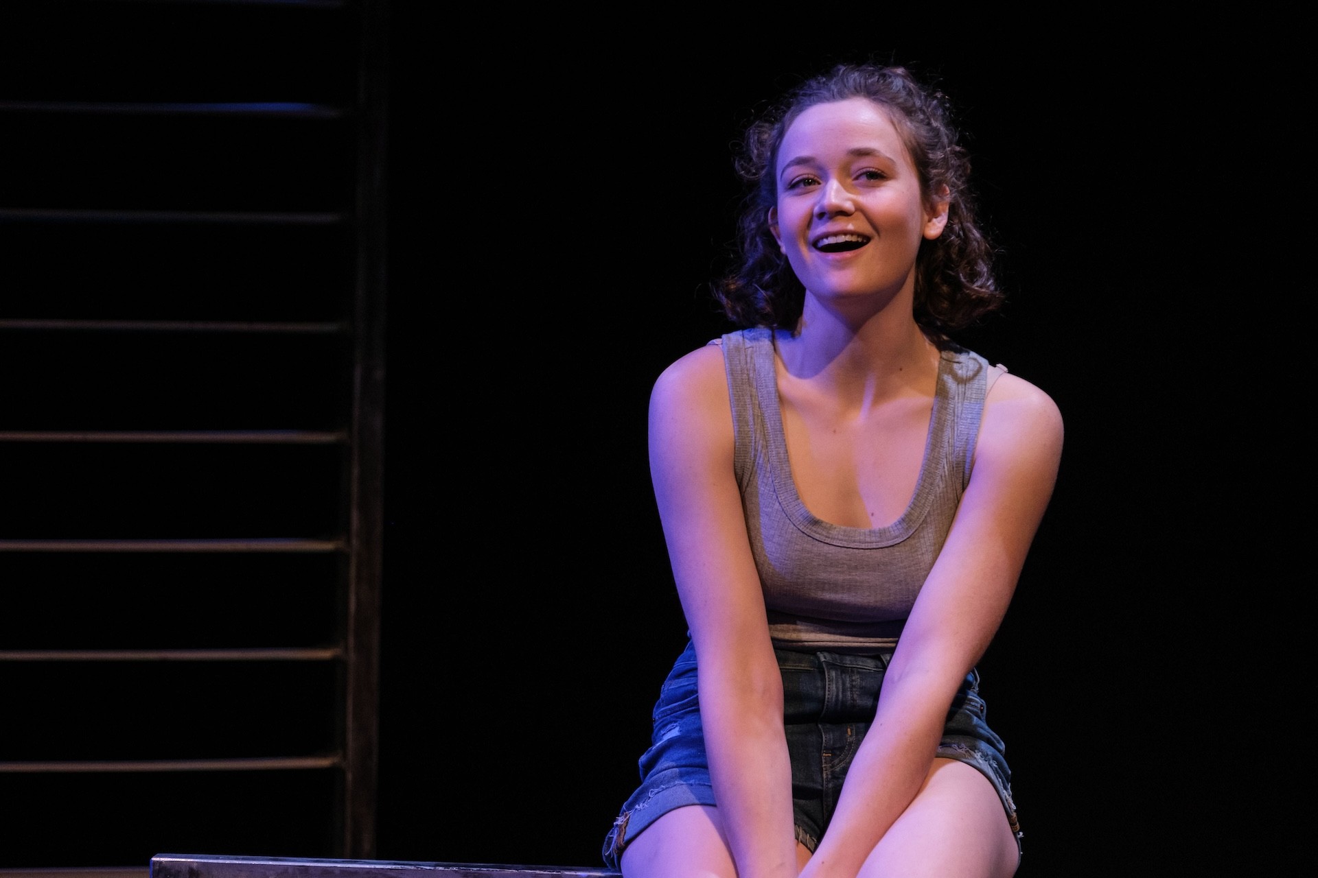 A young performer sits on a platform, smiling softly under gentle stage lighting, wearing a grey tank top and denim shorts against a dark background.