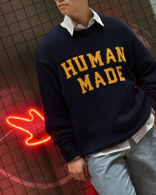 A person wearing a navy sweater with 'HUMAN MADE' written in yellow, posing in front of a grid background with a red neon light shaped like a bird.