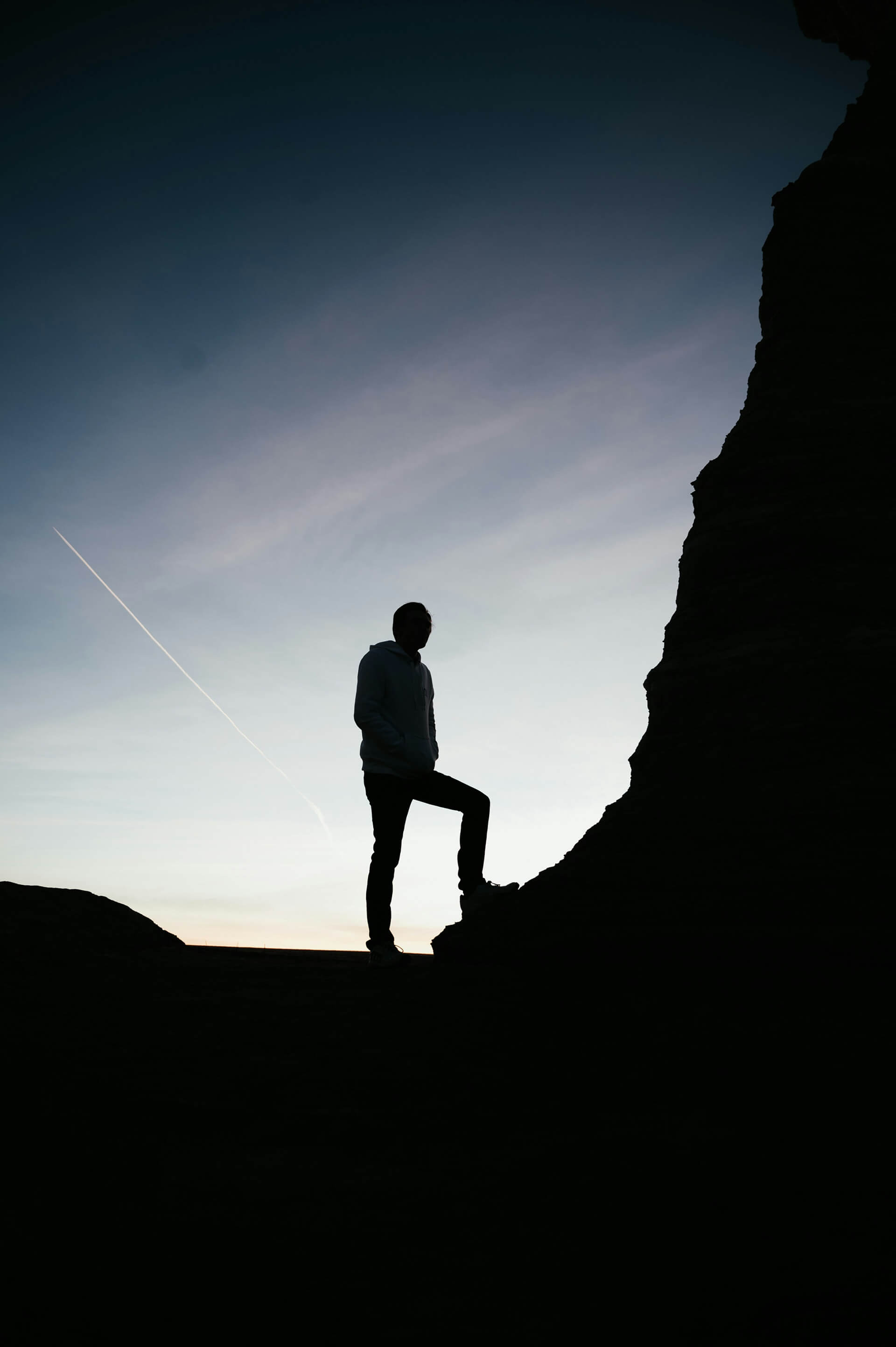 This image features a silhouetted person standing on a rocky terrain at sunset or sunrise. The figure, dressed in a hoodie, stands with one foot elevated on a rock, striking a pose that conveys contemplation or triumph. The background sky transitions from light near the horizon to darker at the top, and a faint airplane contrail cuts diagonally across the sky, adding a sense of motion to the otherwise still scene. The dramatic contrast and composition evoke themes of solitude, achievement, and reflection in nature.