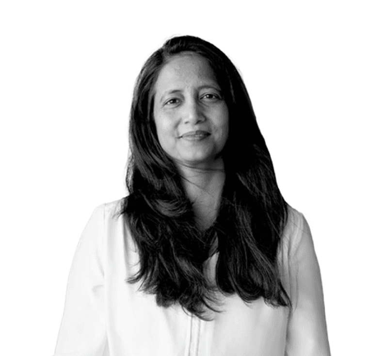 A focused woman with long red hair is seated at a desk, wearing a black blazer and dark blouse. She intently looks at camera, smiling slightly, with an open laptop in front of her. The setting is a warm and serene office setup in the background. Soft daylight filters through, creating a calm atmosphere. The composition captures her in a frontal profile with a clear emphasis on her concentration and the serene indoor setting.