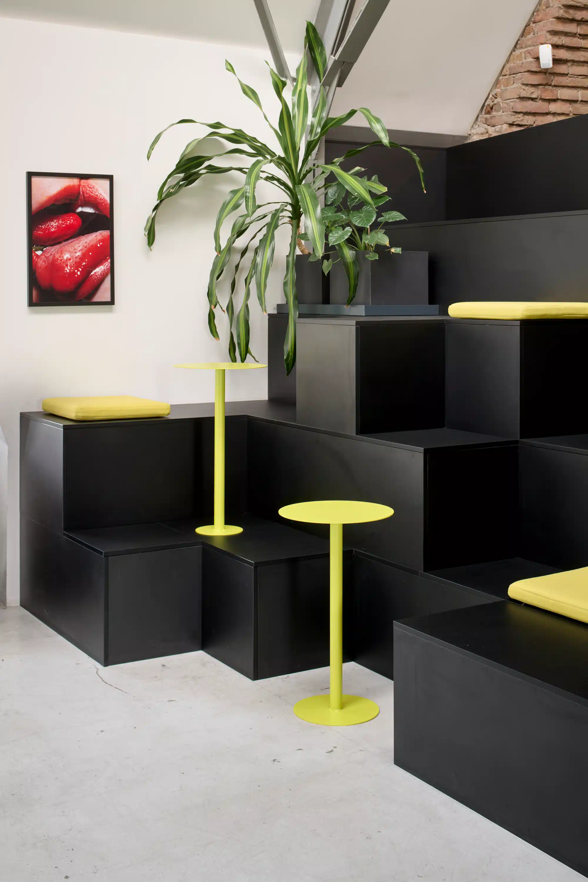 Dojo Office – seating area with black shelf steps, yellow standing tables, indoor plant and wall artwork