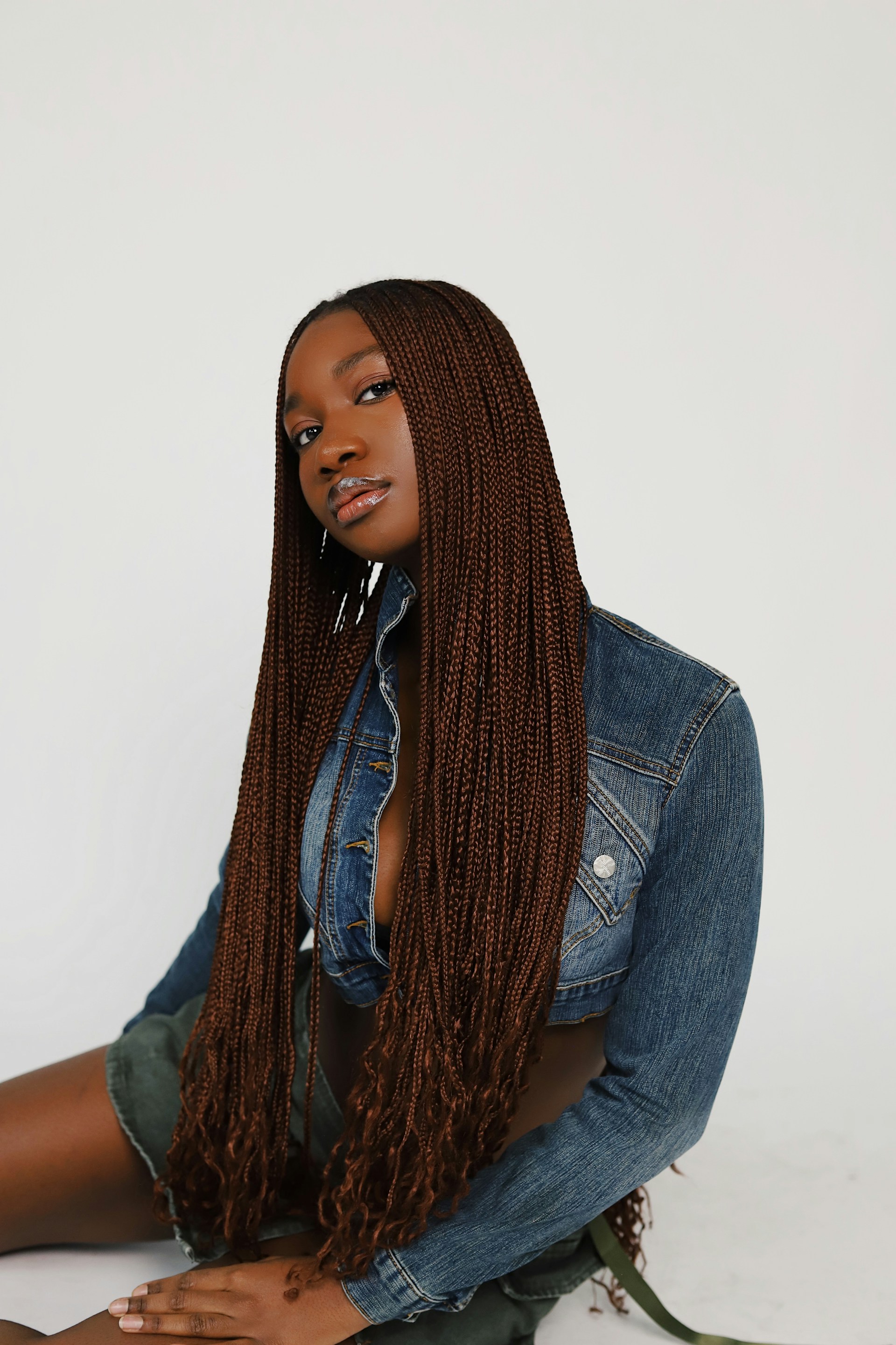 A portrait of a woman with long, reddish-brown micro-braids sitting against a white background. She is wearing a cropped denim jacket and a green utility skirt, looking directly at the camera with a neutral expression.