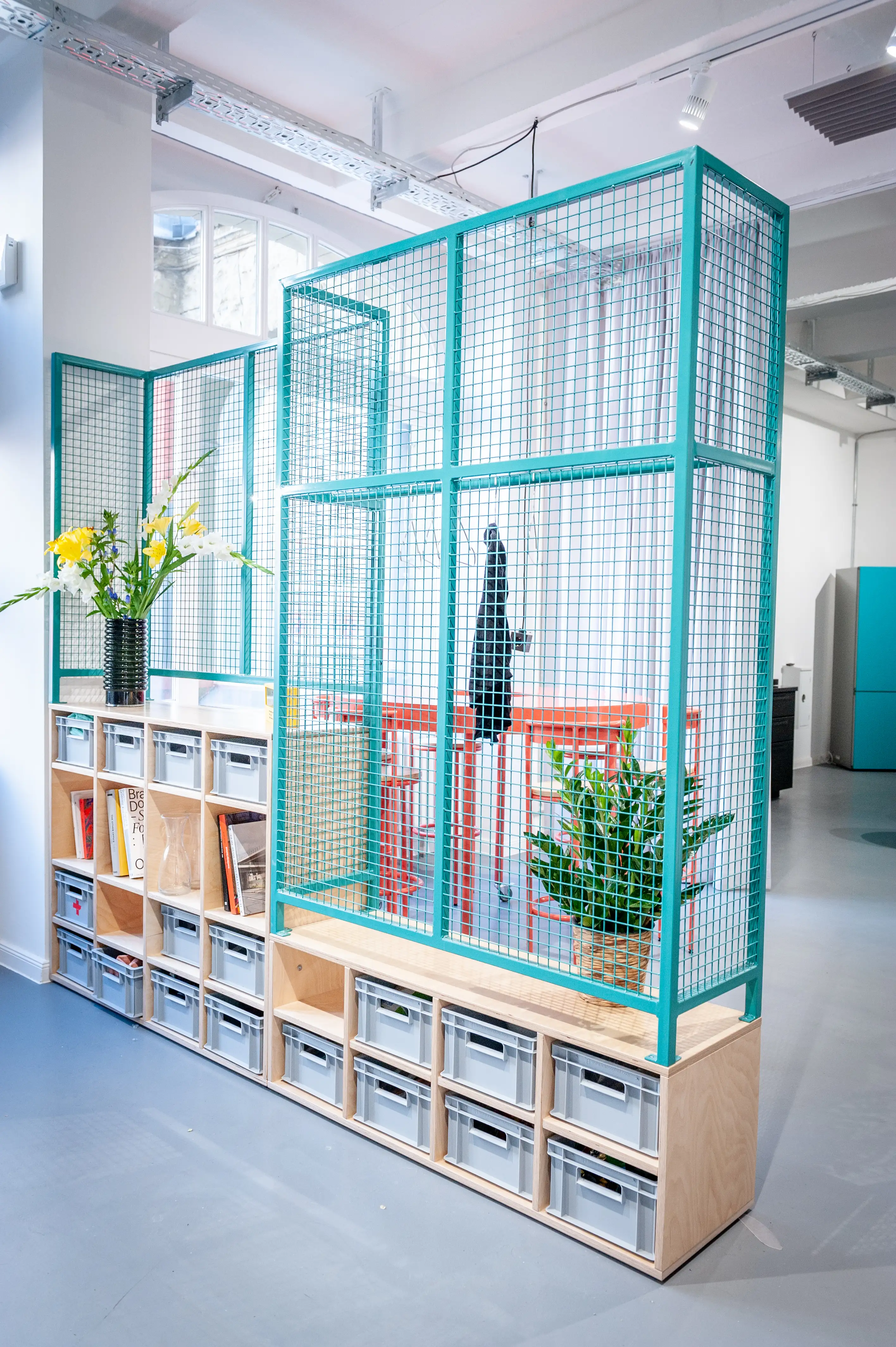 Tall turquoise wire mesh divider with plywood storage shelf at HEY CONTENT STUDIO, with grey crates, plants and a view into the red high-table area behind.