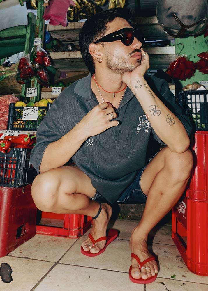 A man wearing a dark gray polo shirt, denim shorts, and red flip-flops crouches in a market setting. He is wearing black sunglasses, a red cord necklace with a silver charm, and has visible tattoos on his arms.