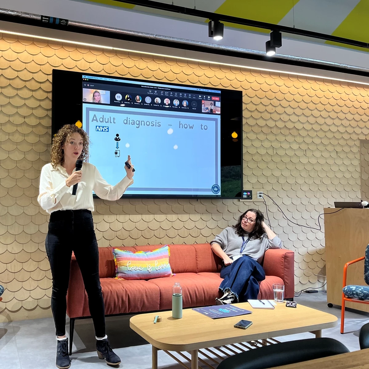 A woman in black jeans and a white shirt stands in front of a screen talking about Adult Diagnosis of ADHD, behind her is a sofa with a woman with a grey jumper sitting on it