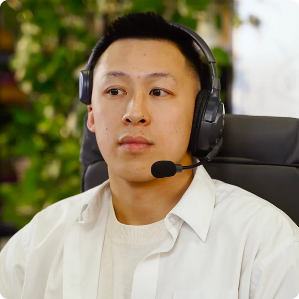 Young man wearing black over-ear headset with microphone, seated in a chair, with blurred greenery in the background.