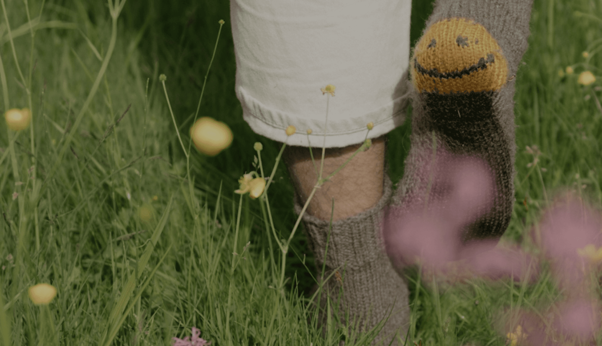 A person's lower body and feet standing in a grassy field filled with yellow and purple flowers, with a knitted sock or leg warmer featuring a smiley face design, creating a serene and whimsical visual composition.