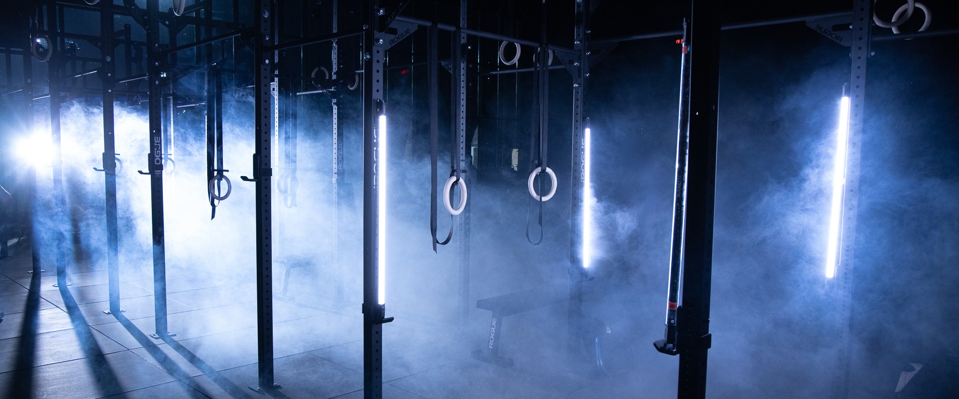 An empty CrossFit gym in cool blue haze, rigs and gymnastic rings lit by vertical strip lights.