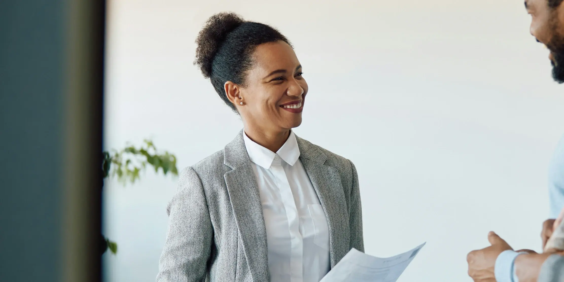 Professional woman in a gray blazer and white collared shirt smiling warmly while holding documents during a conversation with a client