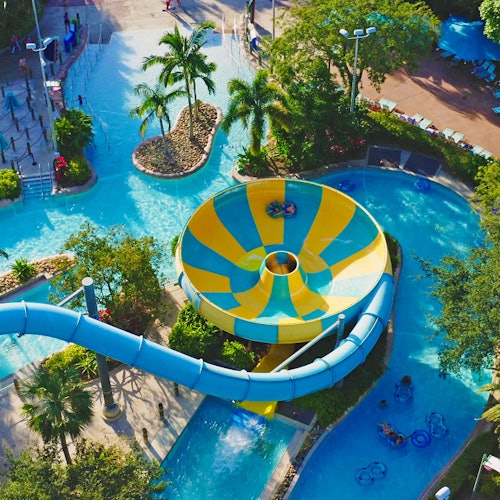 Въздушен изглед на воден парк с голяма жълто-синя фуния и пързалка, заобиколен от дървета и мързелива река.