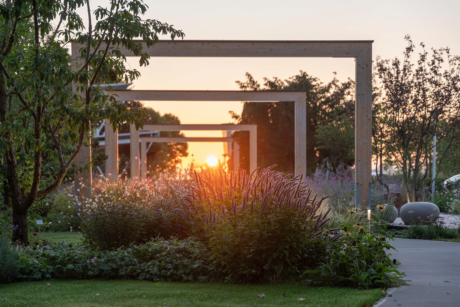 Schaugarten mit Staudenbeeten, Holzrahmen und tief stehender Sonne entlang eines geschwungenen Weges.