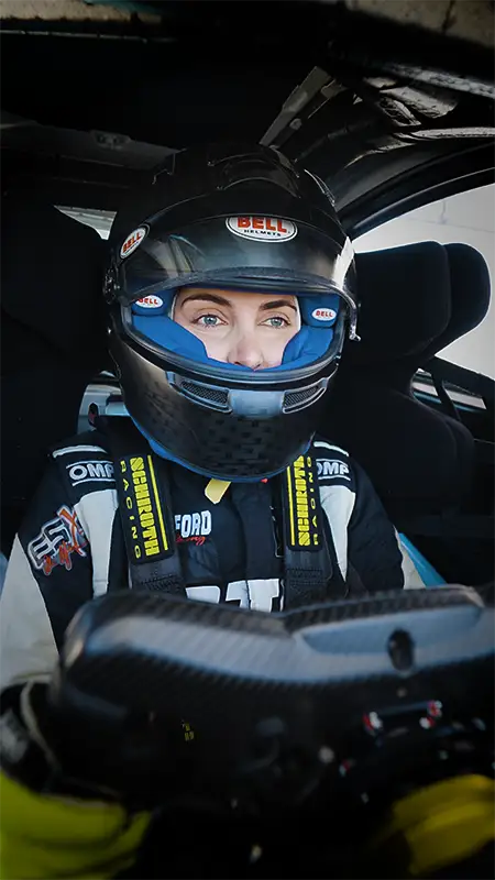 Emelia Hartford wearing a helmet and safety gear inside a race car, focusing intently ahead.