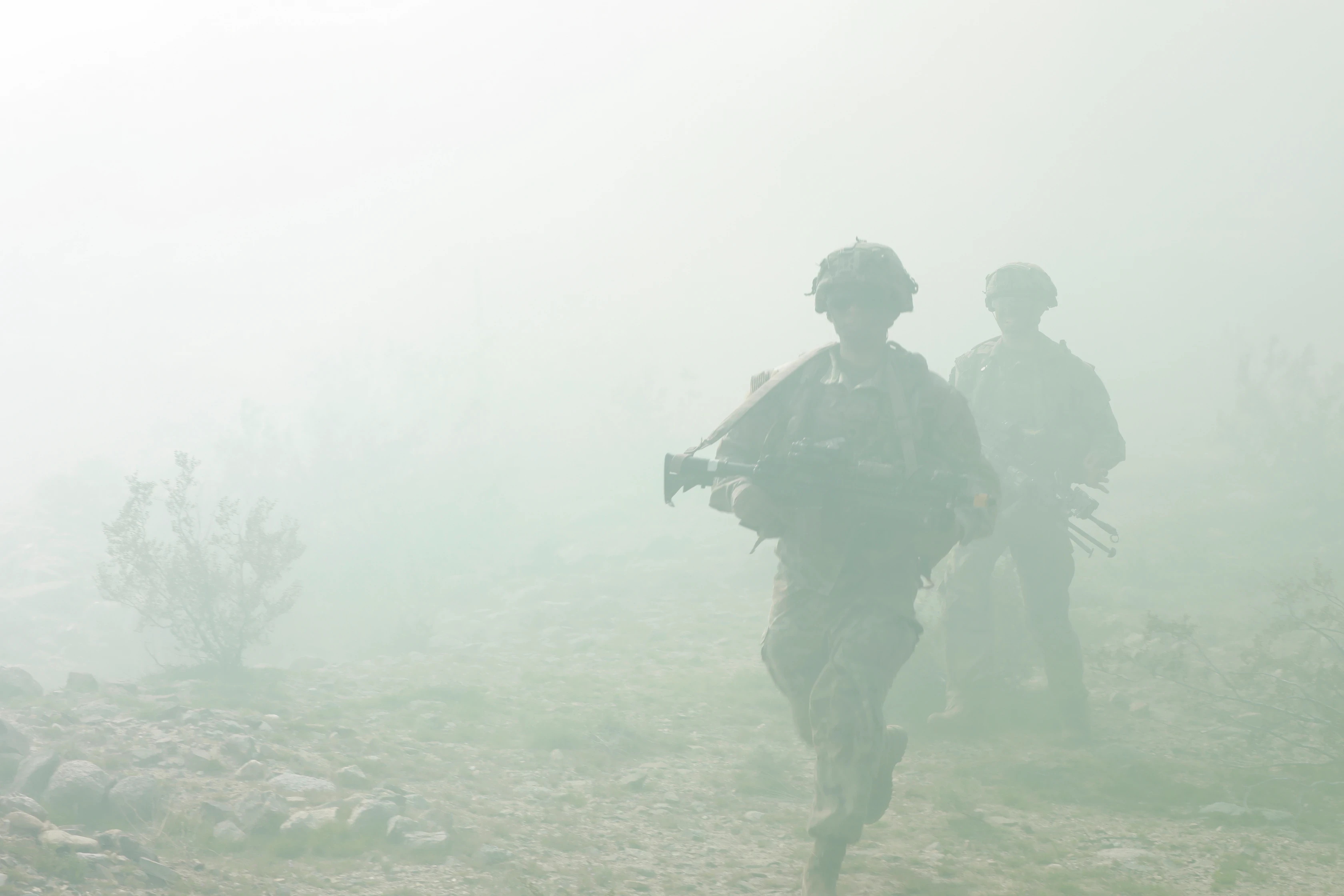 Soldiers in camouflage uniforms moving through a foggy or smoke-filled environment on rocky terrain.