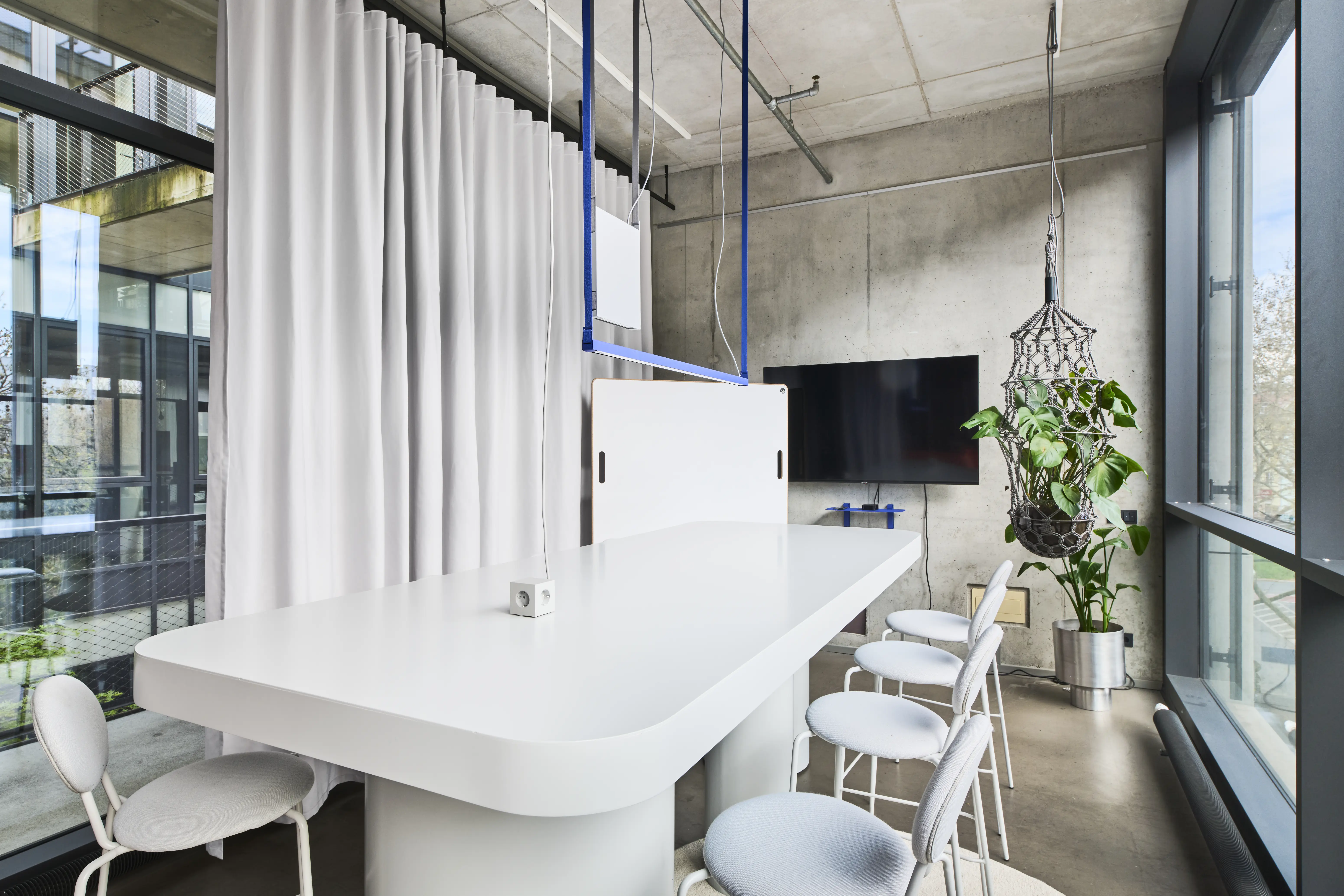 Meeting area in the UNDPLUS SHOWROOM with a rounded white high table, light grey stools, wall screen, hanging plant in a macramé net and white curtain divider.