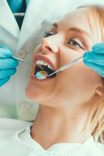 Eine Frau auf einem Zahnarztstuhl lächelt, während ein Zahnarzt ihre Zähne mit zahnärztlichen Instrumenten untersucht.