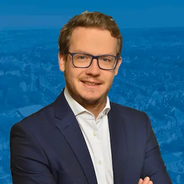 Professional headshot of man with glasses and beard wearing navy suit jacket and white shirt, smiling at camera against blue background with faded cityscape
