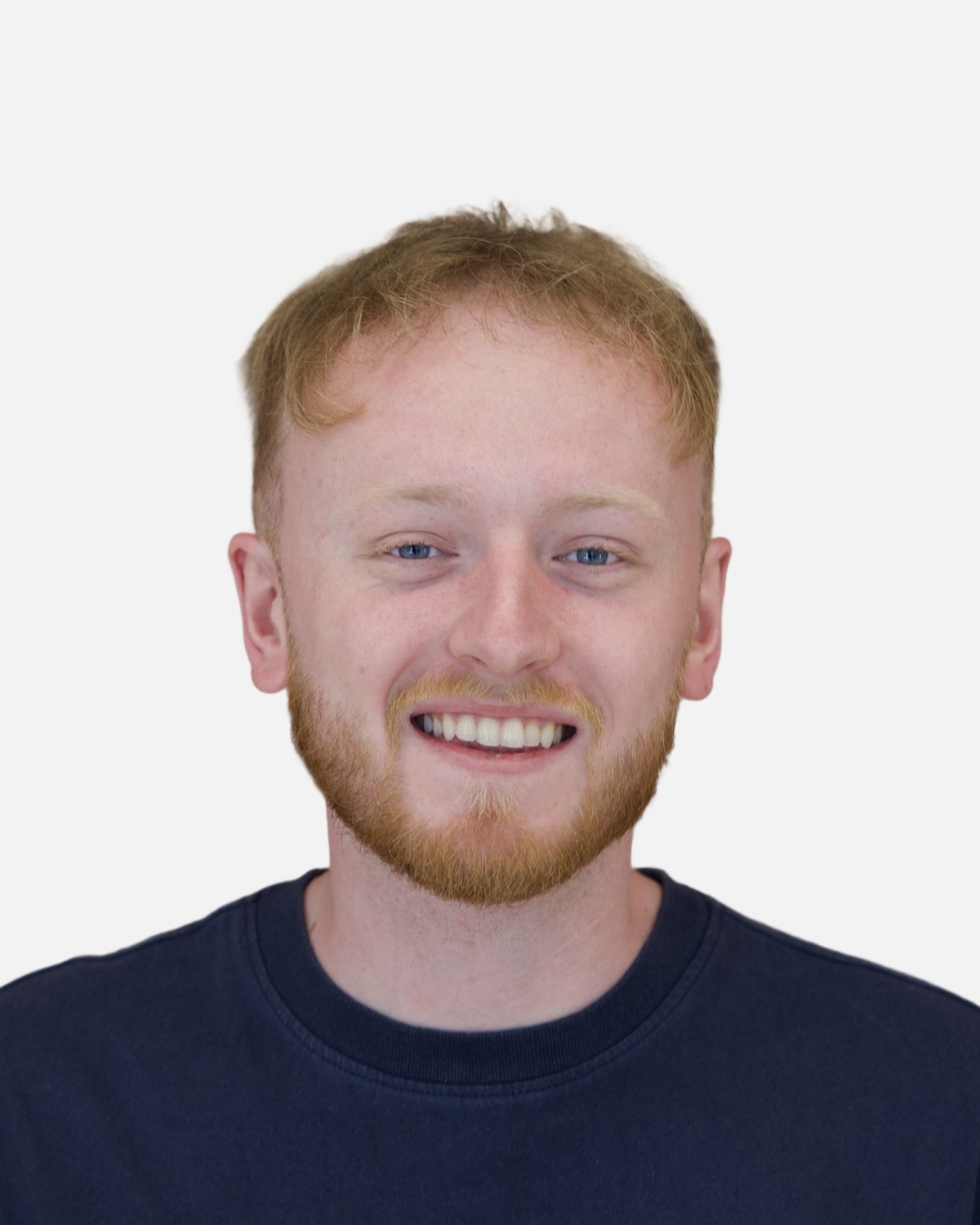 the portrait photo of a young and good-looking man, looking directly to the camera. This man has ginger hair and beard, wears navy shirt on and smiling