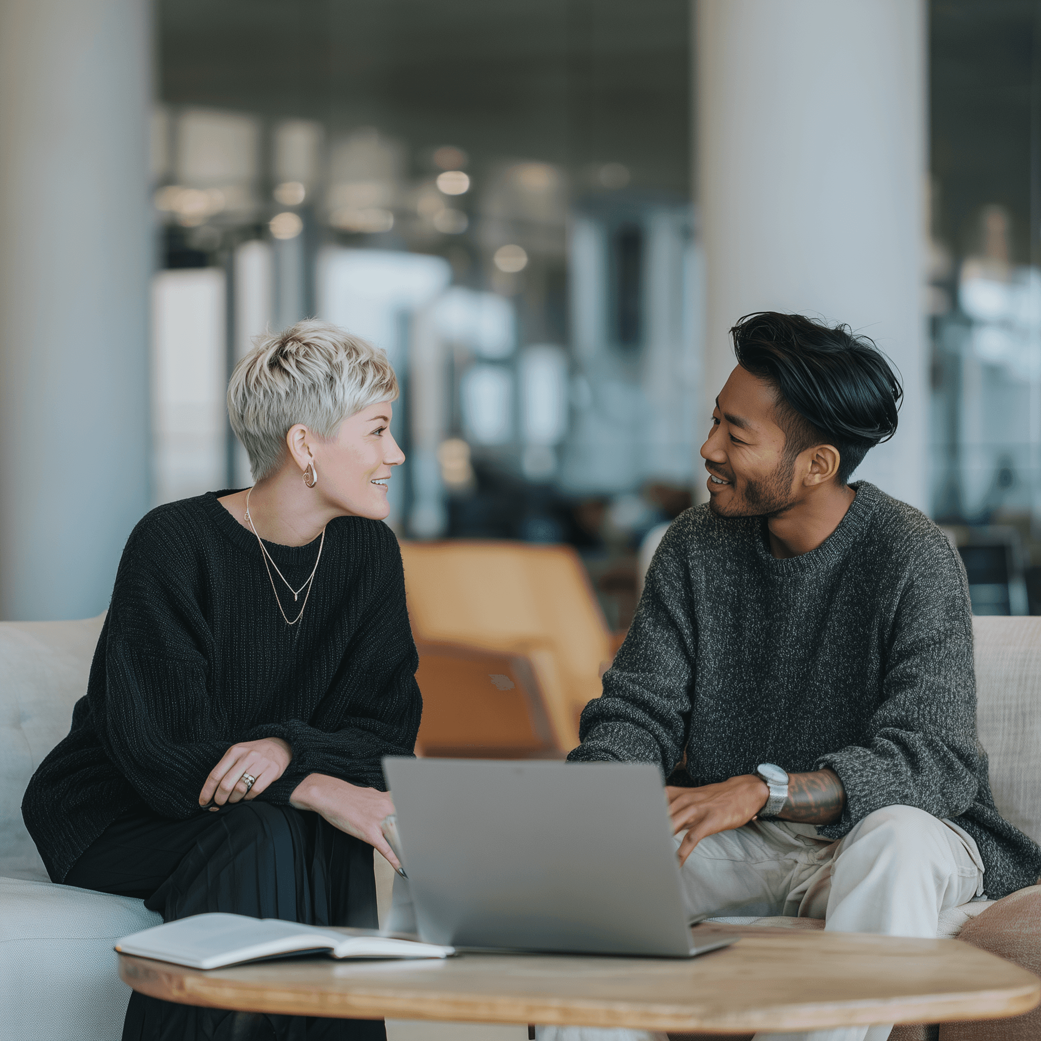 A trendy and cozy work environment where two people chat over a laptop and notebook.