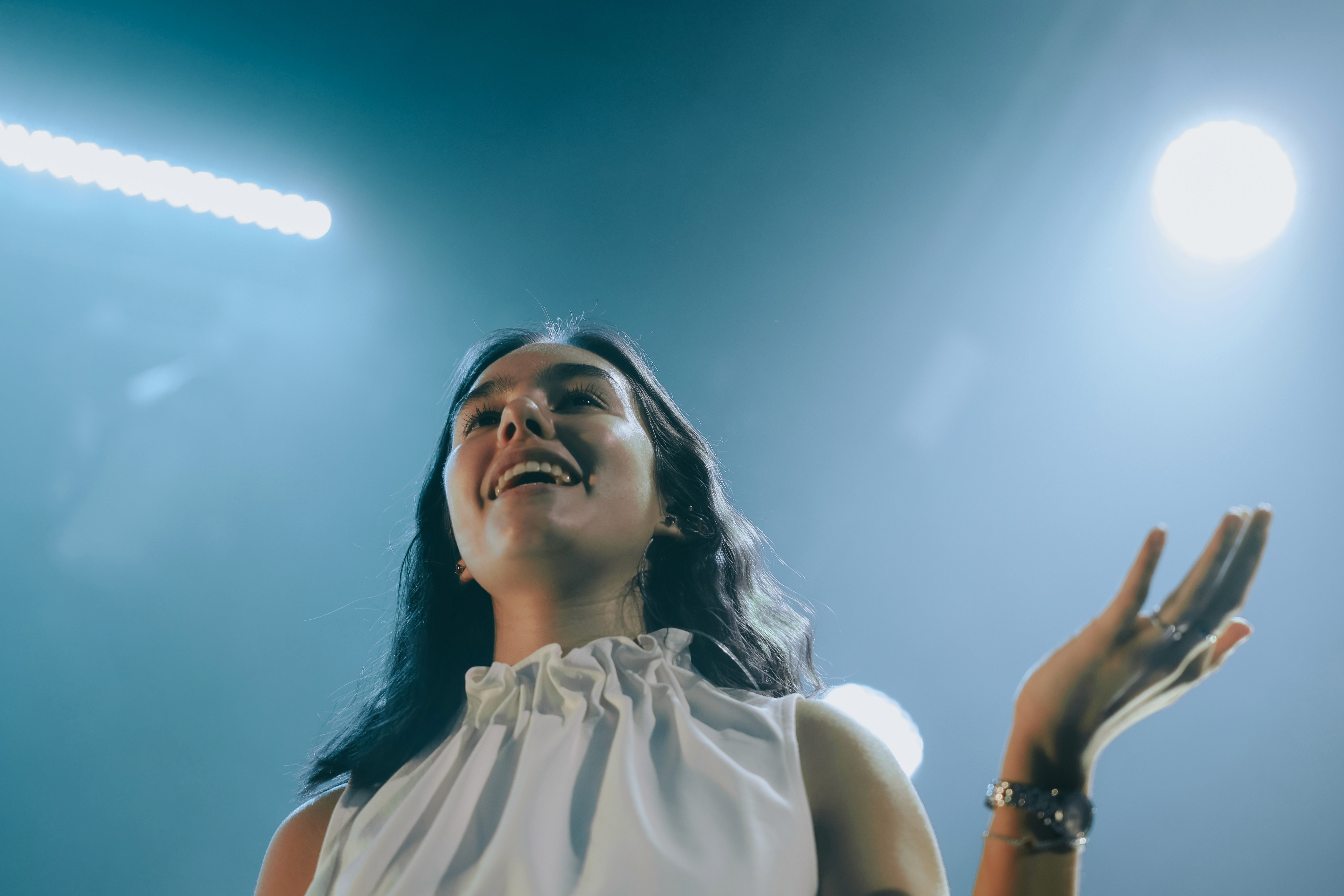 A performer on stage gestures with one hand, surrounded by blue lighting and silhouettes of the audience.