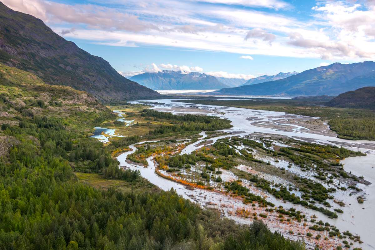 A wide river flows through a green valley toward snow-capped mountains under a blue sky with white clouds.