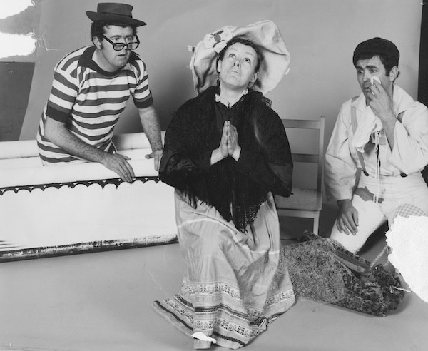 A group portrait of three young adult theatre artists standing and sitting against a dark teal backdrop. A person wearing a navy top and red skirt is seated on a stool in the centre, while two others stand on either side, dressed casually in jeans and T-shirts. All three face the camera with a relaxed, confident expression.