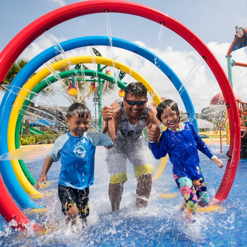 A man and two children run through water jets under colorful hoops at a water park, all wearing swimwear and smiling.