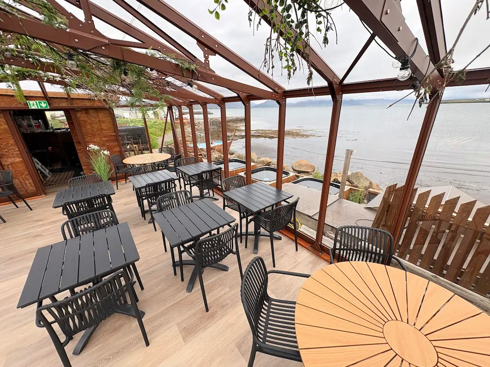 Outdoor seaside cafe seating area with tables and umbrellas overlooking the water at sunset.