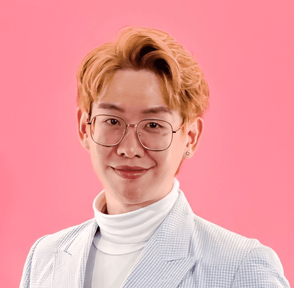Young person with wavy hair poses confidently in sunlight against a modern building backdrop. Wears a black blazer and silver necklace, conveying elegance.
